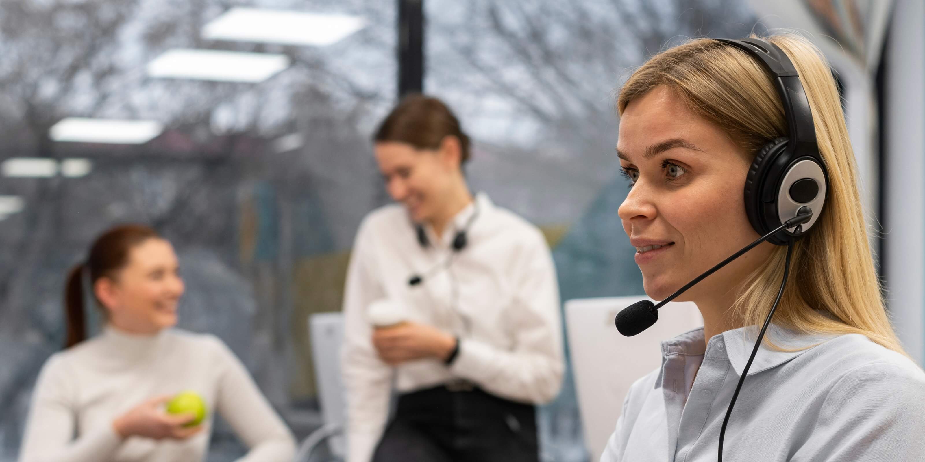 Woman wearing a headset in an office setting with colleagues in the background.